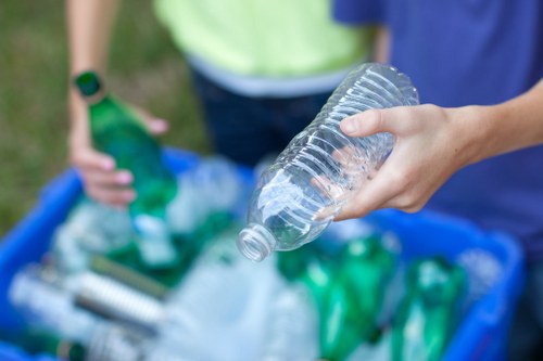 A close-up of a person's hand holding a clear plastic bottle, with a background showing another individual wearing a yellow top and a green wristband, standing outdoors on a grassy area. In the foreground, a blue plastic container filled with various recycling bottles and plastics is visible, highlighting different shapes and shades of green and transparent materials. The setting suggests a waste collection or rubbish removal activity, with the focus on the bottle and the container, emphasizing environmental waste disposal practices typical of local rubbish removal services in Uxbridge or surrounding areas. The image captures the textures of the plastic bottles and the casual outdoor environment, aligning with professional waste management operations by Business Waste Removal Uxbridge.