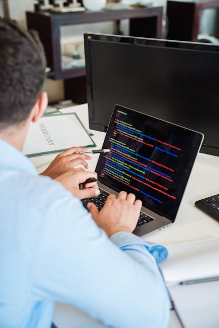 A person seated at a desk working on a laptop computer, with the screen displaying lines of colorful code against a dark background. The individual is using a computer mouse with their right hand and 