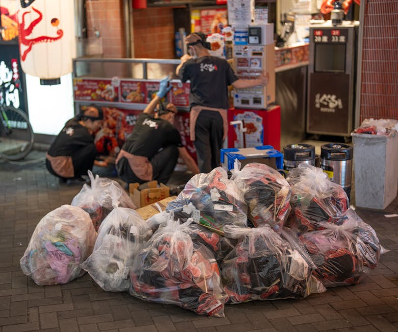 A person wearing a casual plaid shirt with shades of yellow, blue, and gray, along with green gloves, is holding open a large black trash bag made of shiny plastic material. The bag appears to be part