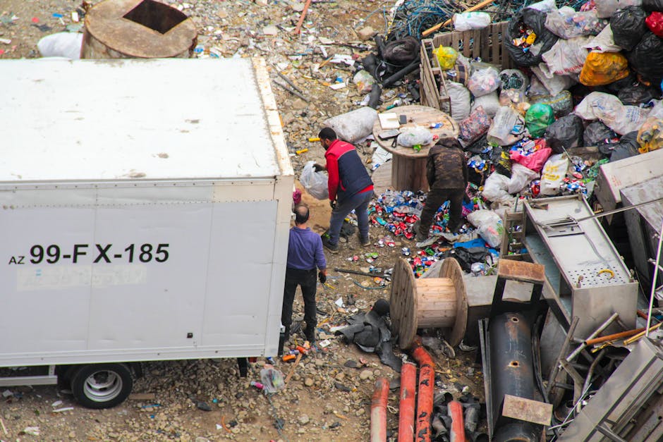 A large pile of mixed household rubbish and waste materials is gathered on a paved street corner in front of a row of commercial storefronts and a building under construction with blue scaffolding. Th